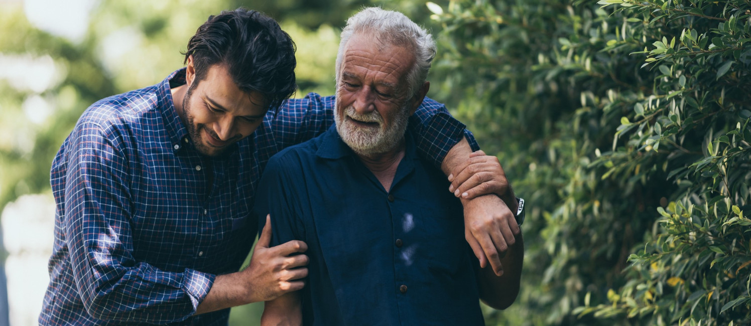 A son helping his elderly father to walk in a bright green park.